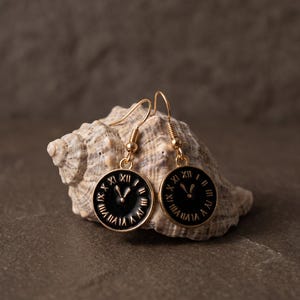 Può includere: Un paio di orecchini pendenti color oro con ciondoli circolari neri. Ogni ciondolo presenta numeri romani dorati e lancette dell'orologio al centro. Gli orecchini sono fissati a ganci dorati per lobi forati.