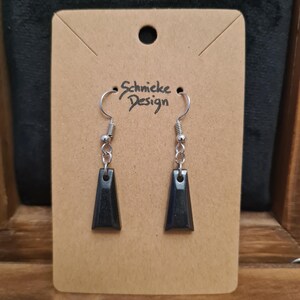 Könnte beinhalten: Ein Paar schwarze Ohrhänger mit silbernen Haken. Die Ohrringe sind dreieckig geformt und haben eine glänzende, glatte Oberfläche. Die Ohrringe befinden sich auf einer braunen Karte mit dem aufgedruckten Text "Schnicke Design".