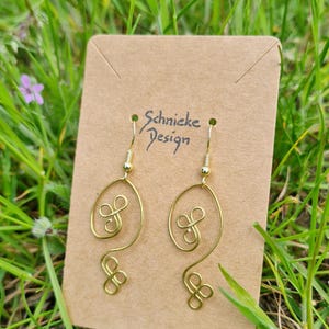 Könnte beinhalten: Ein Paar goldfarbene Drahtohrringe mit einem einzigartigen, fließenden Design. Die Ohrringe werden auf einer braunen Karte mit dem Text "Schnicke Design" präsentiert. Sie haben eine geschwungene ovale Form mit dekorativen Wirbeln.