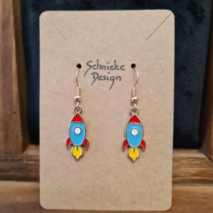Può includere: Un paio di orecchini a forma di razzo dorati con smalto blu, rosso e giallo. Gli orecchini sono su una carta marrone con il testo "Schnieke Design".