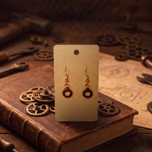 Peut inclure: Une paire de boucles d'oreilles en forme d'engrenage couleur cuivre suspendues à une carte de présentation rectangulaire beige. Chaque boucle d'oreille comporte une petite breloque en forme de roue dentée fixée à un crochet métallique simple.