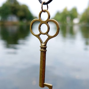 Op de afbeelding: Een gouden sleutel hanger ketting met een decoratief, versierd sleutel ontwerp. De sleutel heeft een ronde, bloemvormige bovenkant en een lange, rechte schacht. De ketting hangt tegen een wazige achtergrond van water en bomen.