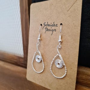 Könnte beinhalten: Silberne tropfenförmige Ohrringe mit einem spiralförmigen Drahtdesign in der Mitte. Die Ohrringe befinden sich auf einer braunen Karte mit dem Text "Schmieke Design".