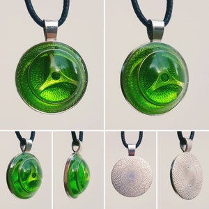 Könnte beinhalten: Eine grüne Halskette mit einem runden Anhänger mit geometrischem Muster. Der Anhänger ist rund und hat eine silberne Öse. Die Halskette besteht aus schwarzem Band.