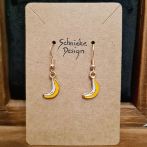 Könnte beinhalten: Ein Paar goldfarbene Hängeohrringe mit Bananen-Design. Jeder Ohrring hat eine gelbe Banane mit weißem Streifen und goldfarbenen Haken. Die Ohrringe werden auf einer braunen Karte mit dem Text "Schmicke Design" präsentiert.