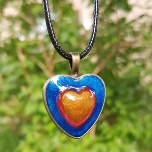 Pode incluir: Um colar com um pingente em forma de coração com um design de esmalte azul, vermelho e dourado. O pingente está em um cordão preto.