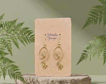 Puede incluir: Un par de pendientes de alambre en tono dorado con un diseño abstracto y en espiral. Los pendientes están sujetos a una tarjeta de presentación rectangular de color tostado con el texto Schnieke Design. El alambre forma un bucle continuo.
