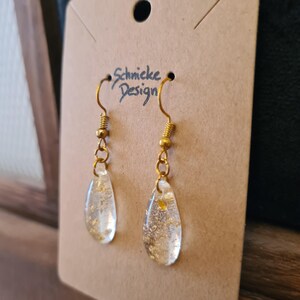 Puede incluir: Pendientes de aro dorados con resina transparente en forma de lágrima que contiene escamas doradas. Los pendientes están en una tarjeta marrón con el texto "Schnicke Design".
