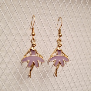 Puede incluir: Un par de pendientes de aro dorados con un diseño de bailarina y una falda de color lavanda. La bailarina está delineada en oro.