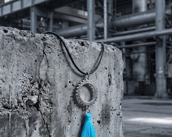 Peut inclure: Un collier composé d'un cordon noir, d'un pendentif en métal argenté en forme d'engrenage et d'un pompon en fil bleu cyan vif suspendu au bas de l'engrenage. Le pendentif est attaché au cordon qui repose sur une surface grise texturée.