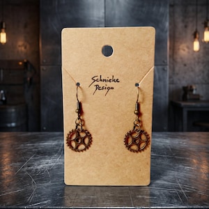 Peut inclure: Une paire de boucles d'oreilles pendantes en forme d'engrenage couleur bronze avec des crochets, présentées sur un support en carton beige rectangulaire portant le texte imprimé Schnicke Design. Les boucles présentent un motif de roue dentée.