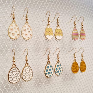 Può includere: Una collezione di orecchini a tema pasquale con ganci color oro. Gli orecchini presentano ciondoli a forma di uovo con vari disegni, tra cui cuori, pois e strisce, in colori come bianco, giallo e blu. Gli orecchini sono esposti su uno sfondo bianco.