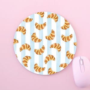 Könnte beinhalten: Rundes Mauspad mit hellblau-weiß gestreiftem Hintergrund und einem Muster aus Croissants. Eine rosa Computermaus ist zu sehen. Ein dekoratives und funktionelles Schreibtischzubehör.