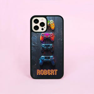 May include: Black phone case featuring a design of four colorful game controllers stacked vertically. The name "ROBERT" is displayed in gold lettering at the bottom. The case has a black border and is set against a pink background.
