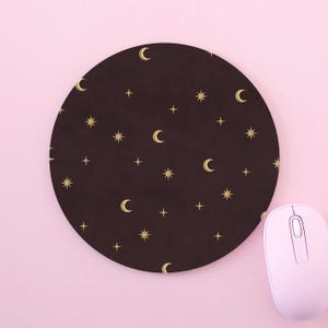 May include: A circular mouse pad featuring a dark brown background with a repeating pattern of gold-colored crescent moons and various star shapes. The mouse pad is positioned next to a light pink computer mouse on a matching pink surface.