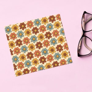 Może przedstawiać: Kwadratowa, wzorzysta ściereczka do czyszczenia z powtarzającym się wzorem uśmiechniętych kwiatów w odcieniach brązu, żółci, pomarańczy i błękitu na kremowym tle. Okulary z ciemną oprawką spoczywają na różowej powierzchni.