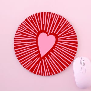 May include: A circular red mouse mat featuring a pink heart design at its centre, encircled by radiating lines. A white computer mouse is positioned to the right, set against a pink backdrop.
