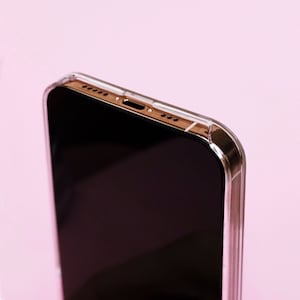 May include: A close-up of a black smartphone in a clear case. The phone is on a pink background.