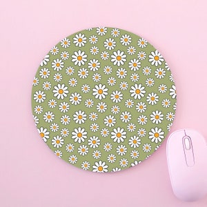 Pode incluir: Tapete de rato redondo com fundo verde coberto por margaridas brancas com centros amarelos. Um rato de computador rosa claro está ao lado do tapete numa superfície rosa. Acessório de computador.
