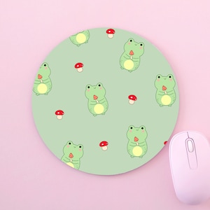 May include: A round mouse mat with a repeating pattern of green frogs holding red mushrooms. The background is a light green colour.