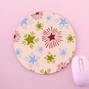 May include: A round mouse pad featuring a light pink background with a repeating pattern of stars in red, olive green, and light blue. Each star is surrounded by radiating lines, creating a burst effect across the surface of the circular pad.