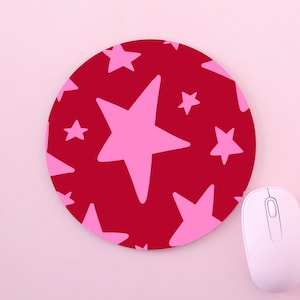 May include: A circular mouse mat featuring a red backdrop adorned with pink stars of different sizes. A white computer mouse is placed to the right of the mat on a pink surface. The mouse mat is designed for computer use.