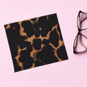 May include: A square microfiber cleaning cloth with a tortoiseshell pattern, next to a pair of eyeglasses with a brown frame. The cloth features a repeating pattern of dark brown shapes on a light brown background. The eyeglasses have a light pink background.