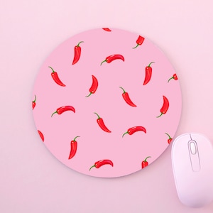 Op de afbeelding: Een ronde roze muismat met een herhalend patroon van rode pepers. Een witte draadloze muis ligt op een roze oppervlak naast de muismat.