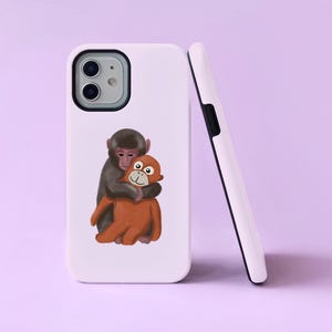 Könnte beinhalten: Weißes Handycase mit einer Cartoon-Illustration von zwei sich umarmenden Affen. Ein Affe ist braun, der andere orange. Ein zweites weißes Handycase lehnt an dem ersten, mit einer schwarzen Kameralinse und schwarzem Rand.