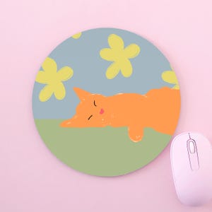 May include: A circular mouse pad featuring a graphic illustration of an orange cat sleeping on a light green surface against a light blue background with scattered yellow flower shapes. A light pink computer mouse sits to the right.