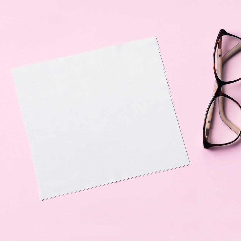 May include: A white microfiber cleaning cloth with a scalloped edge on a pink background. A pair of brown-framed eyeglasses is lying on the right side of the image.