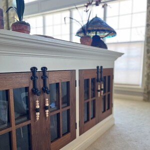 May include: A white cabinet with two wooden doors, each with a black metal handle and a glass pane. The doors are decorated with wooden beads hanging from the handles.