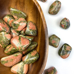 May include: A wooden bowl filled with green and red tumbled stones. The stones are a mix of green and red, with some having a more speckled appearance. The bowl is sitting on a white surface.
