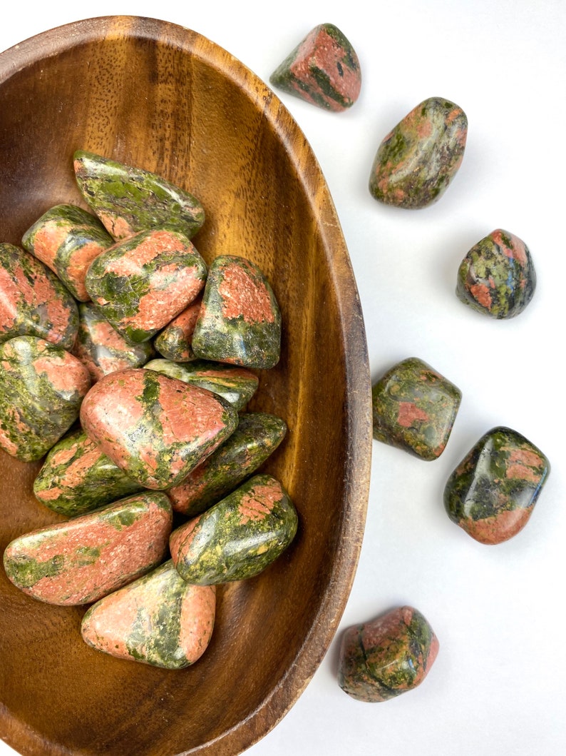 May include: A wooden bowl filled with green and red tumbled stones. The stones are a mix of green and red, with some having a more speckled appearance. The bowl is sitting on a white surface.