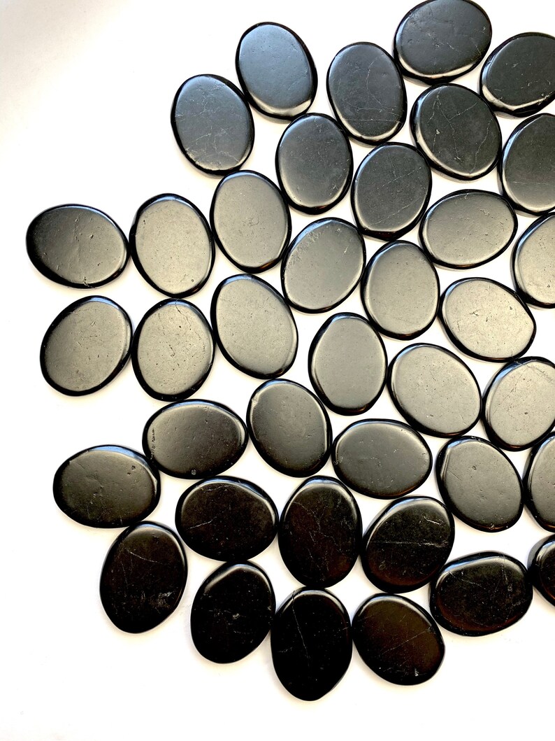 May include: A collection of smooth, black oval-shaped stones. The stones are arranged in a semi-circle pattern on a white background.