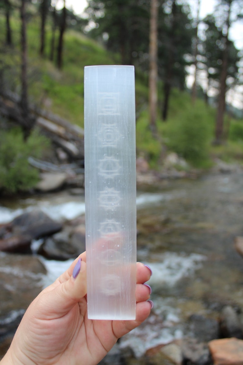Pu&ograve; includere: Una bacchetta di selenite trasparente con una superficie liscia e lucida. La bacchetta &egrave; lunga circa 15 cm e ha una forma rettangolare. La bacchetta &egrave; tenuta in una mano su uno sfondo sfocato di un ruscello e alberi.