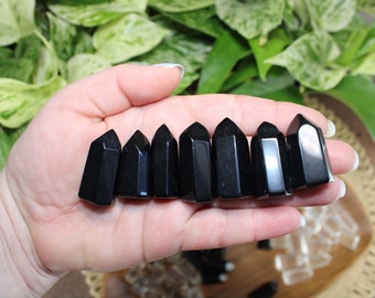 May include: Eight black obsidian points, also known as black glass, are arranged in a row on a hand. The points are smooth and polished, with a slight shine.