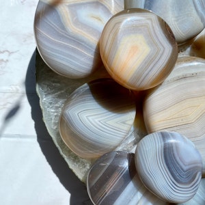 May include: A collection of polished agate stones in various shades of gray and white with intricate banding patterns. The stones are smooth and rounded, and they are arranged in a circular pattern.