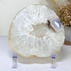 May include: A white agate slice with a natural crystal formation in the center. The slice is on a clear acrylic stand.