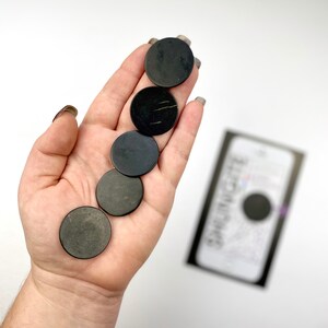 May include: Five black round smooth stones, also known as "worry stones", are held in a hand. The stones are approximately 1 inch in diameter.