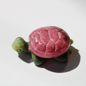 May include: A small, carved turtle figurine. The turtle's shell is a vibrant pink with carved geometric patterns, while the head and legs are a muted green. The figurine is set against a plain white background, casting a shadow.