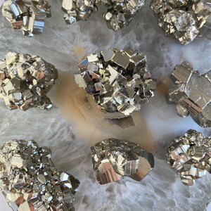 May include: A collection of pyrite crystals, also known as fool's gold, arranged on a white agate slice. The crystals are a metallic gold color and have a variety of shapes and sizes.