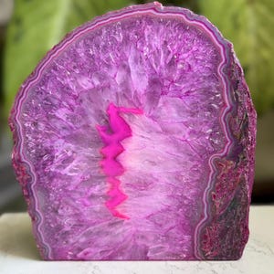 May include: A polished agate slice featuring a vibrant magenta and purple crystalline center with a bright pink jagged core. The outer edge displays concentric bands of white, grey, and deep purple, creating a natural semi-circular stone display.