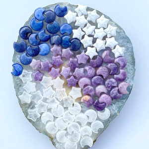 May include: A collection of blue, purple, and clear crystal moon and star shapes on a white agate slice. The crystals are arranged in a circular pattern.