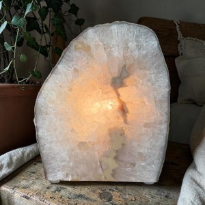 May include: A translucent white and pale pink agate geode lamp with a rough, crystalline texture. The interior of the stone is illuminated from within, casting a warm, soft glow through the jagged, sparkling quartz crystals and mineral layers.