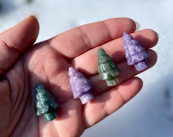 May include: Four small carved crystal trees in different shades of green and purple. The trees are arranged in a row on a hand.