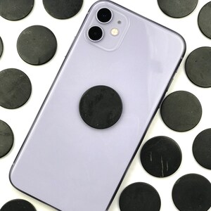 May include: A purple smartphone with a black phone grip and a black camera lens. The phone is surrounded by black round stones on a white background.