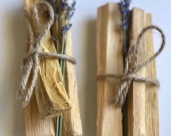 May include: Two bundles of Palo Santo sticks tied with twine and decorated with dried lavender flowers. The sticks are a light brown color and the lavender flowers are a light purple color.