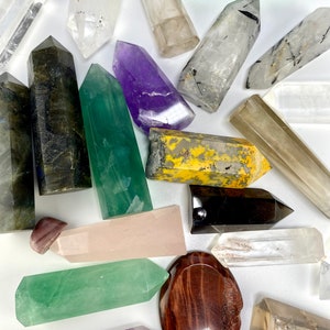 May include: A collection of 16 polished crystal points in various colors and shapes, including clear quartz, amethyst, green fluorite, labradorite, and black obsidian. The crystals are arranged on a white surface.