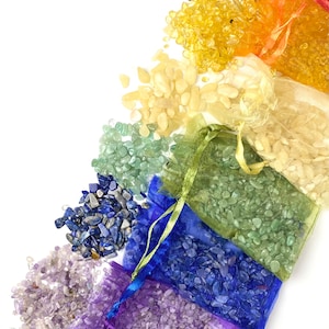 May include: Seven small bags filled with colorful tumbled stones, arranged in a rainbow pattern. The bags are made of sheer fabric and are tied with ribbons. The stones are amethyst, lapis lazuli, green aventurine, citrine, clear quartz, carnelian, and red jasper.